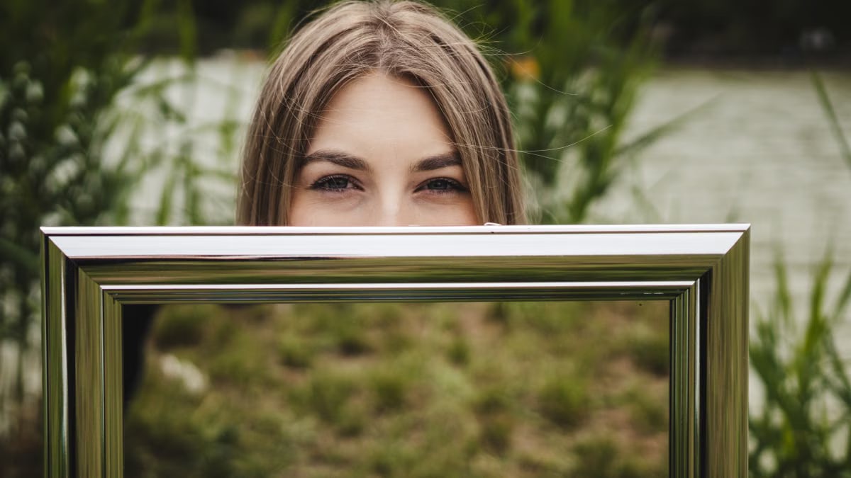 Woman holding a mirror outdoors reflecting grass and nature, showcasing creativity.